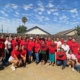 About 30 UFW members and allies pose as a group in Bakersfield, CA. They wear bright red t-shirts and hold flags with the UFW logo, a stylized black eagle with its wings shaped like an inverted Aztec pyramid.