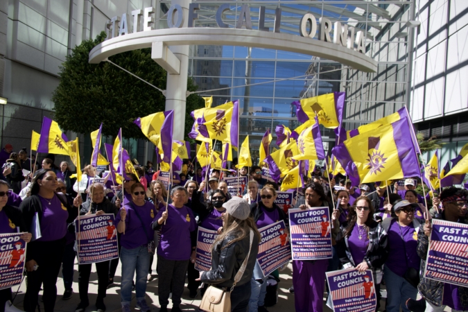 SOTU-Fastfood-Photo A group of 30 or so fast-food workers and allies wearing purple t-shirts gather outside a state building in Oakland, CA. They hold bright flags with the Fast Food Council logo, depicting a worker raising a flag. They hold signs that read "Fast Food Council: Fair wages; Fair working hours; Fair working conditions"