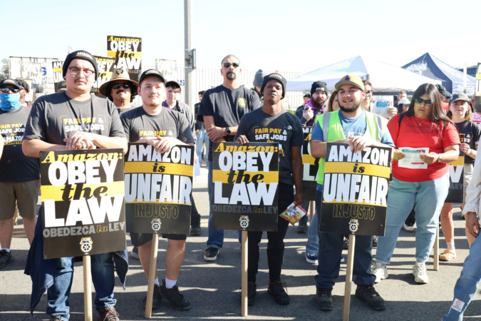 SOTU-Photo-IEwarehouse Workers on strike and allies are pictured on strike outside the Amazon KSBD Air Hub in San Bernardino, CA. Four workers in the foreground hold signs reading "Amazon: Obey the law (Obedezca la Lay)" and "Amazon is Unfair (Injusto)