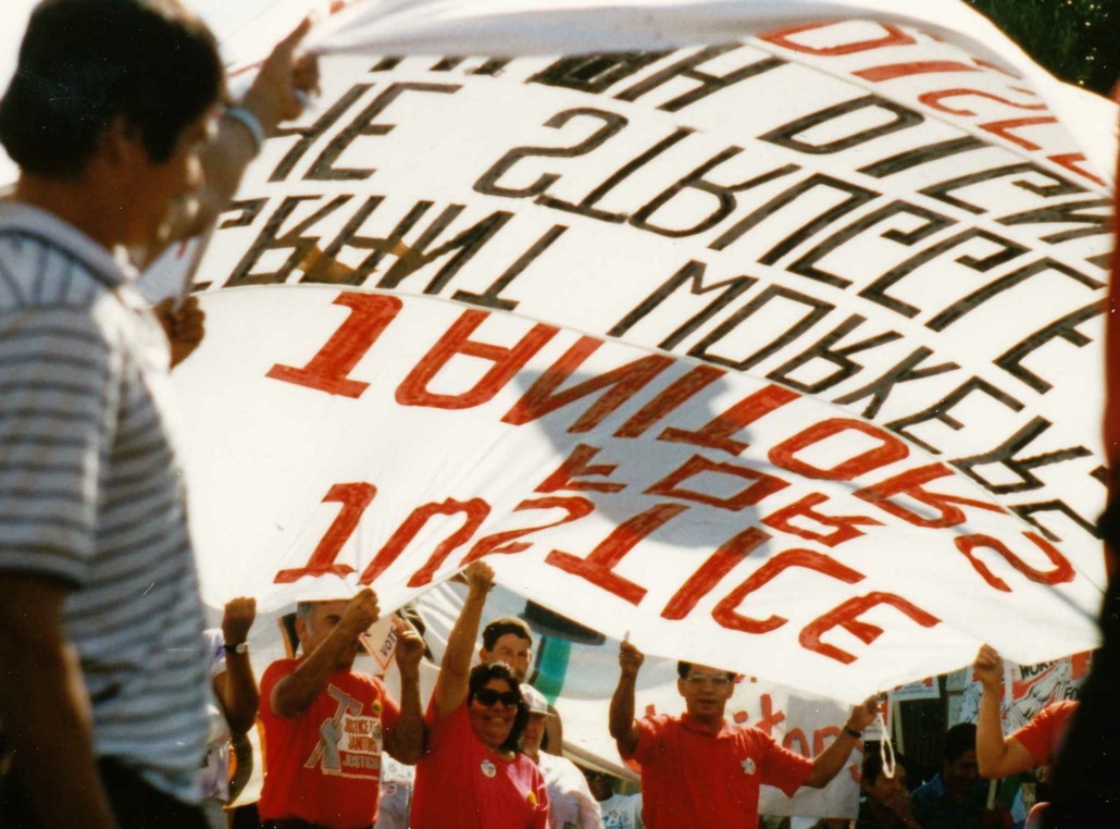 A photo taken from within a janitors union march shows participants lifting a large banner over their heads at a march against Prop. 187 in May, 1994. SEIU-USWW (Justice for Janitors) Collection, IRLE Archives.